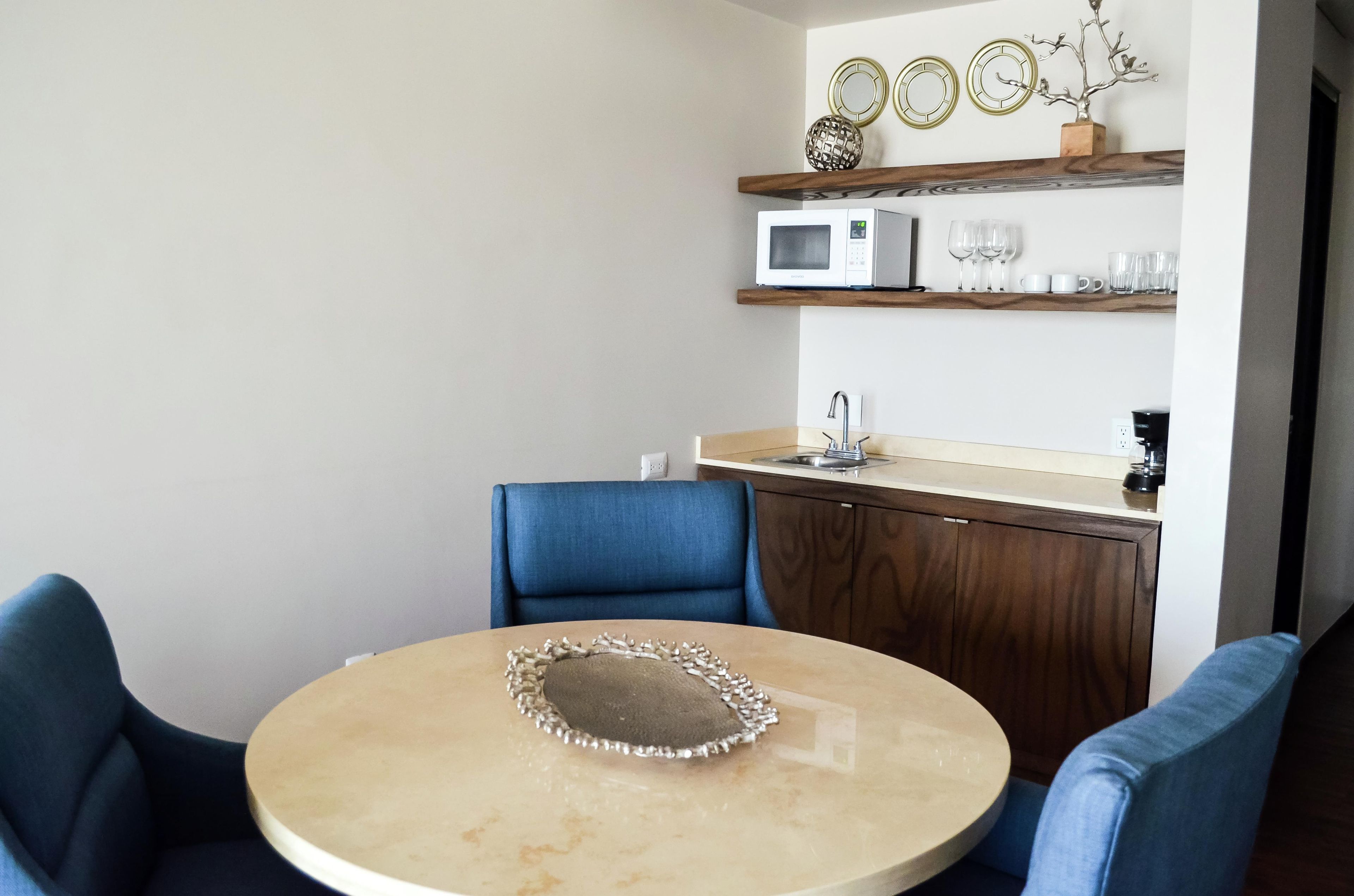 Un petit coin repas avec une table ronde, deux chaises bleues, un four à micro-ondes posé sur une étagère et un meuble avec un évier.