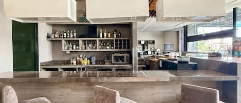 Zona de bar moderna con vigas de madera en el techo, lámparas colgantes y una barra con estantería para vinos. Hay tres taburetes de bar tapizados.