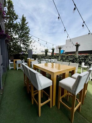 An outdoor seating area with a wooden table and chairs, surrounded by greenery and string lights.