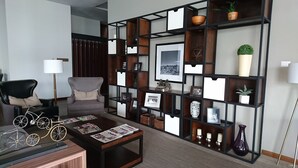Una sala de estar moderna con un sofá negro, una mesa de centro de madera con revistas, una bicicleta decorativa, una estantería negra con diversos objetos y un amplio ventanal con cortinas.
