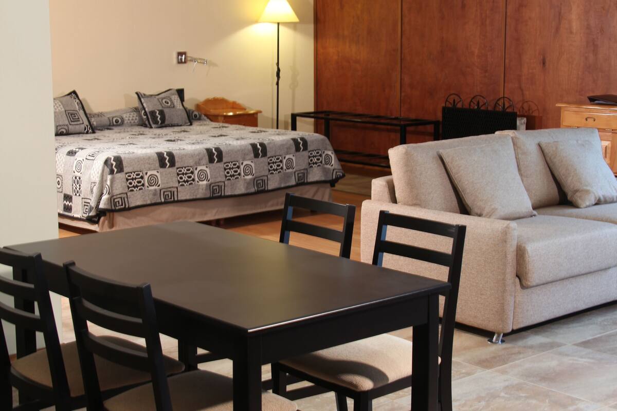 A living room with a sofa, a dining table with chairs, a lamp, and a bed with patterned bedding.