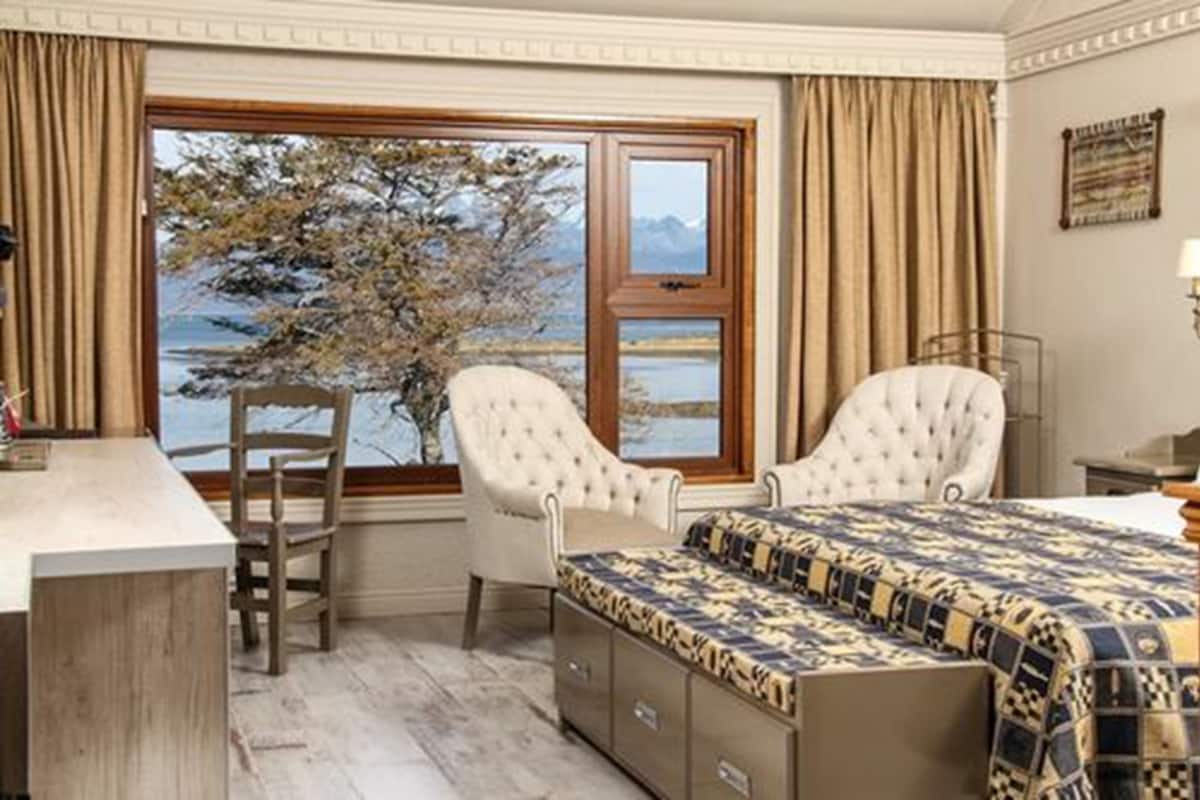 A bedroom with a large window, a bed with a patterned bedspread, two upholstered chairs, and a wooden desk.