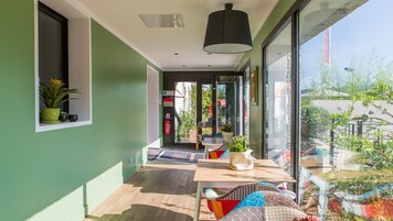 Quarto moderno com uma grande porta de vidro deslizante que dá para uma varanda, piso de madeira e uma mesa com almofadas e um vaso de planta.