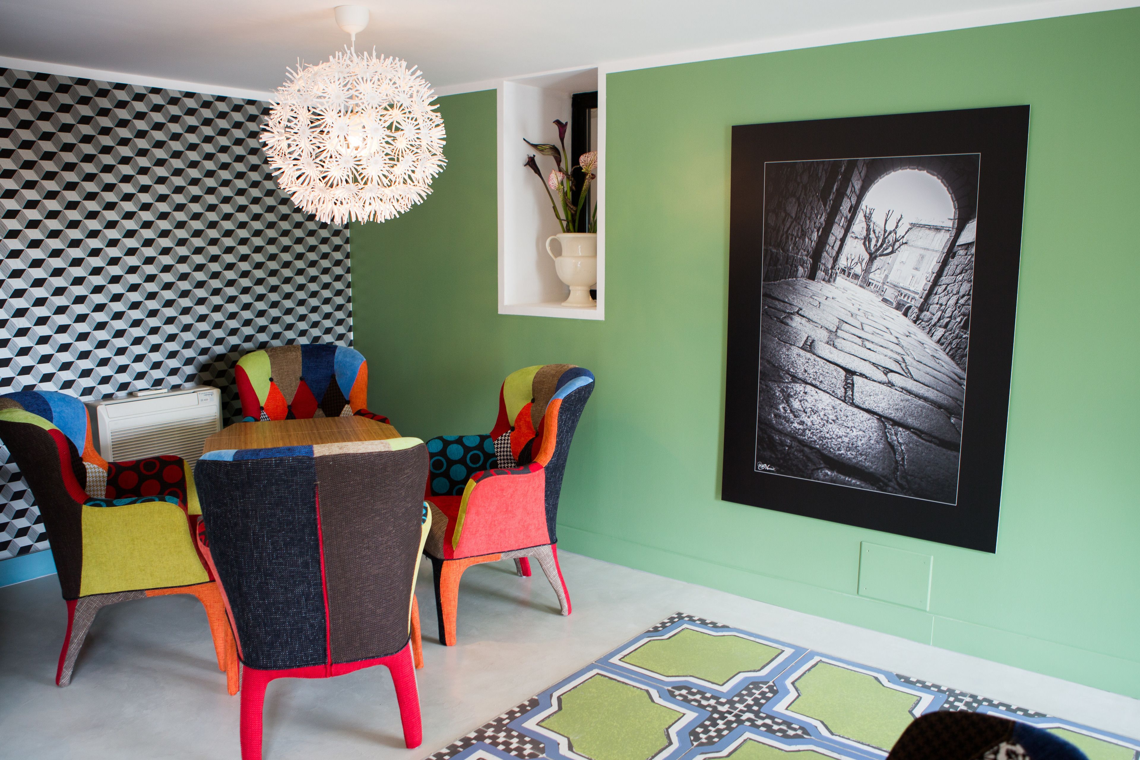 A dining area with a round table and colorful chairs, a pendant light, and a framed black and white photograph on the wall.