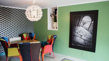 A dining area with a round table and colorful chairs, a pendant light, and a framed black and white photograph on the wall.