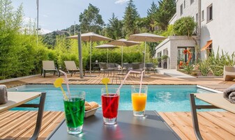 Uma área à beira da piscina com espreguiçadeiras, guarda-sóis e vista para um edifício ao fundo.