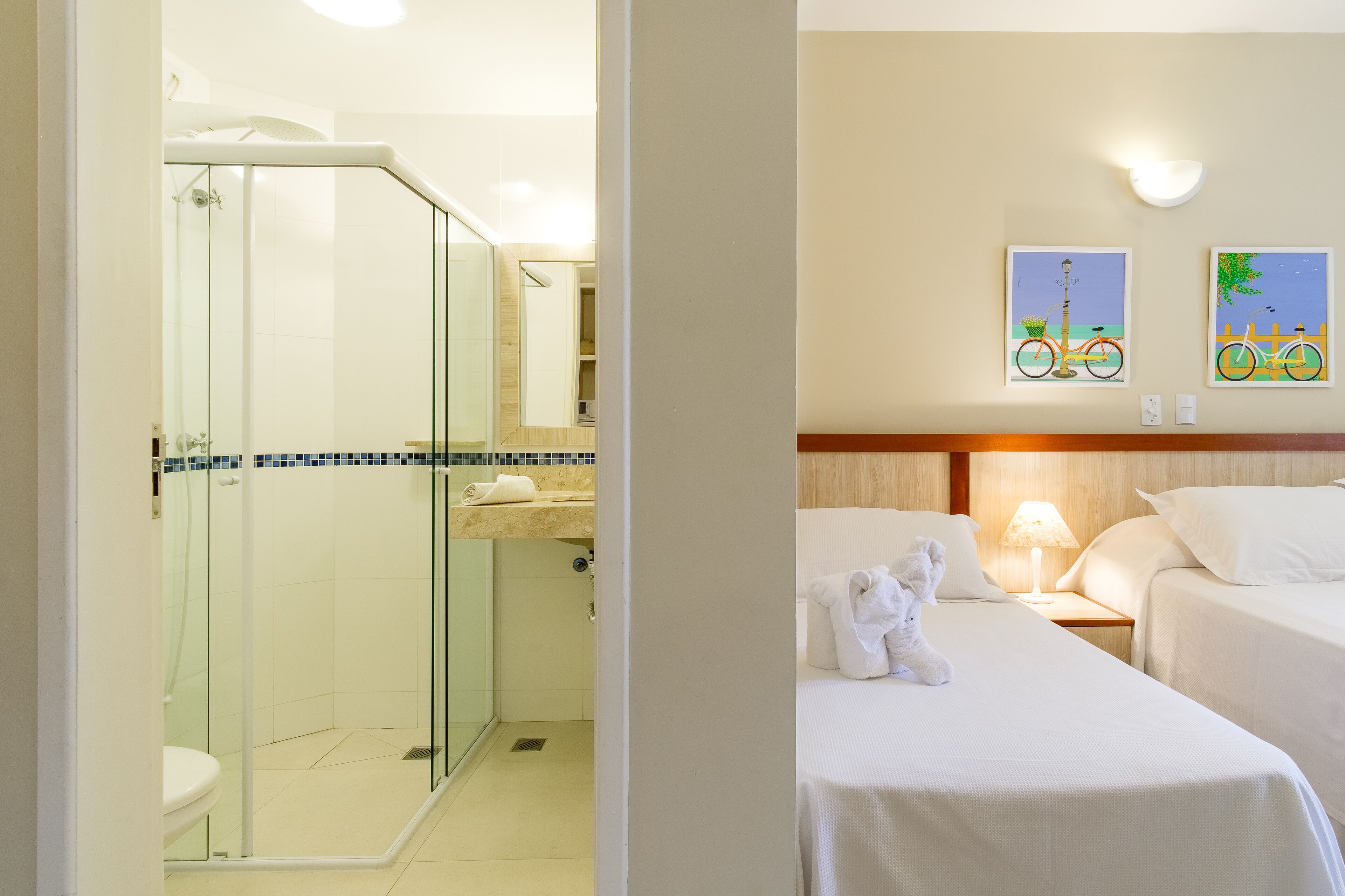 Ein Hotelbad mit einer gläsernen Duschkabine, einer Toilette und einem Waschbecken. Es gibt zwei Betten mit weißer Bettwäsche und einen Teddybären auf einem Bett.
