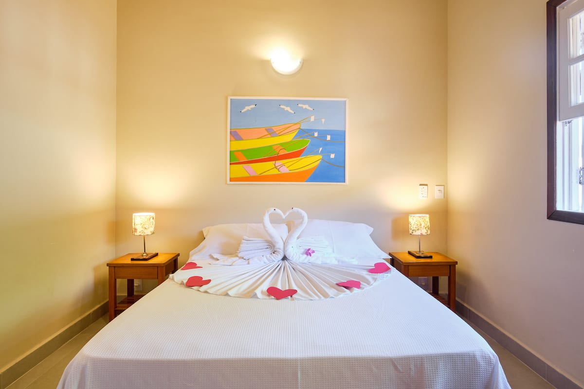 Una cama bien hecha con una toalla dispuesta en forma de corazón, un cuadro de barcos en la pared y dos lámparas de noche.