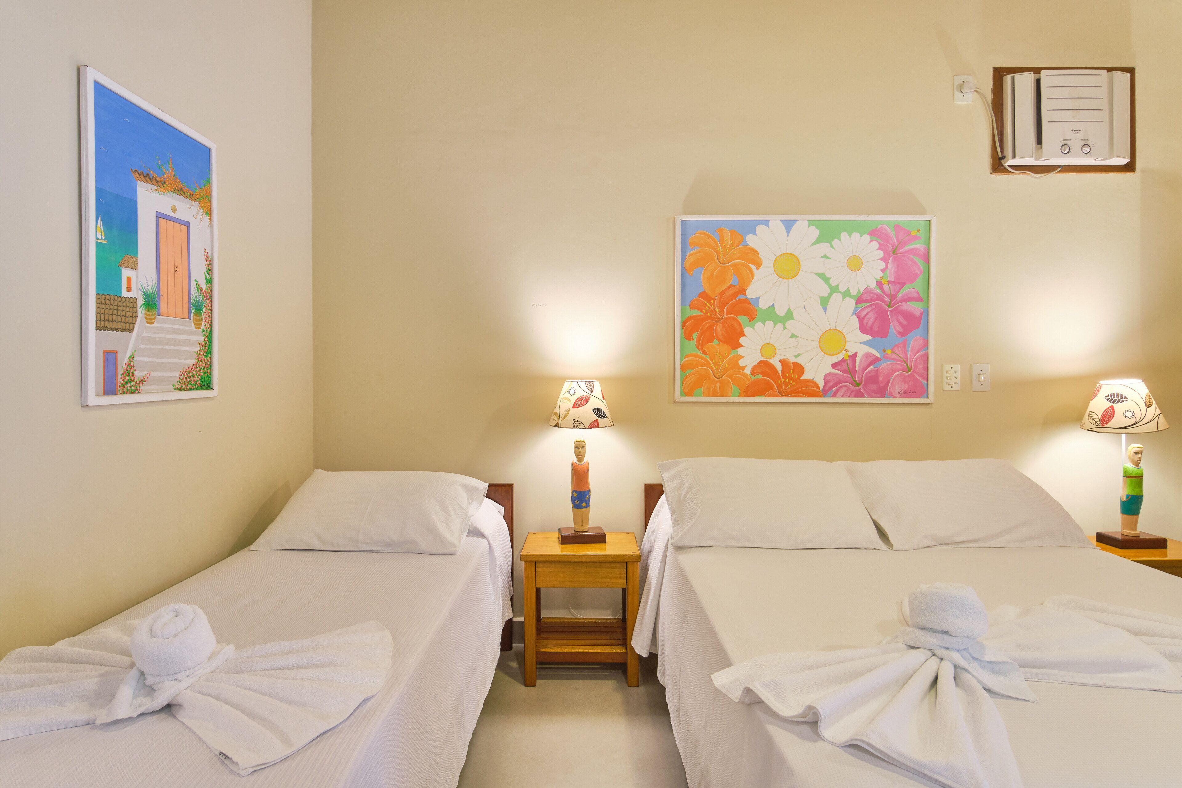 Una habitación de hotel con dos camas, ropa de cama blanca, una mesita de noche de madera con una lámpara y un cuadro de flores en la pared.