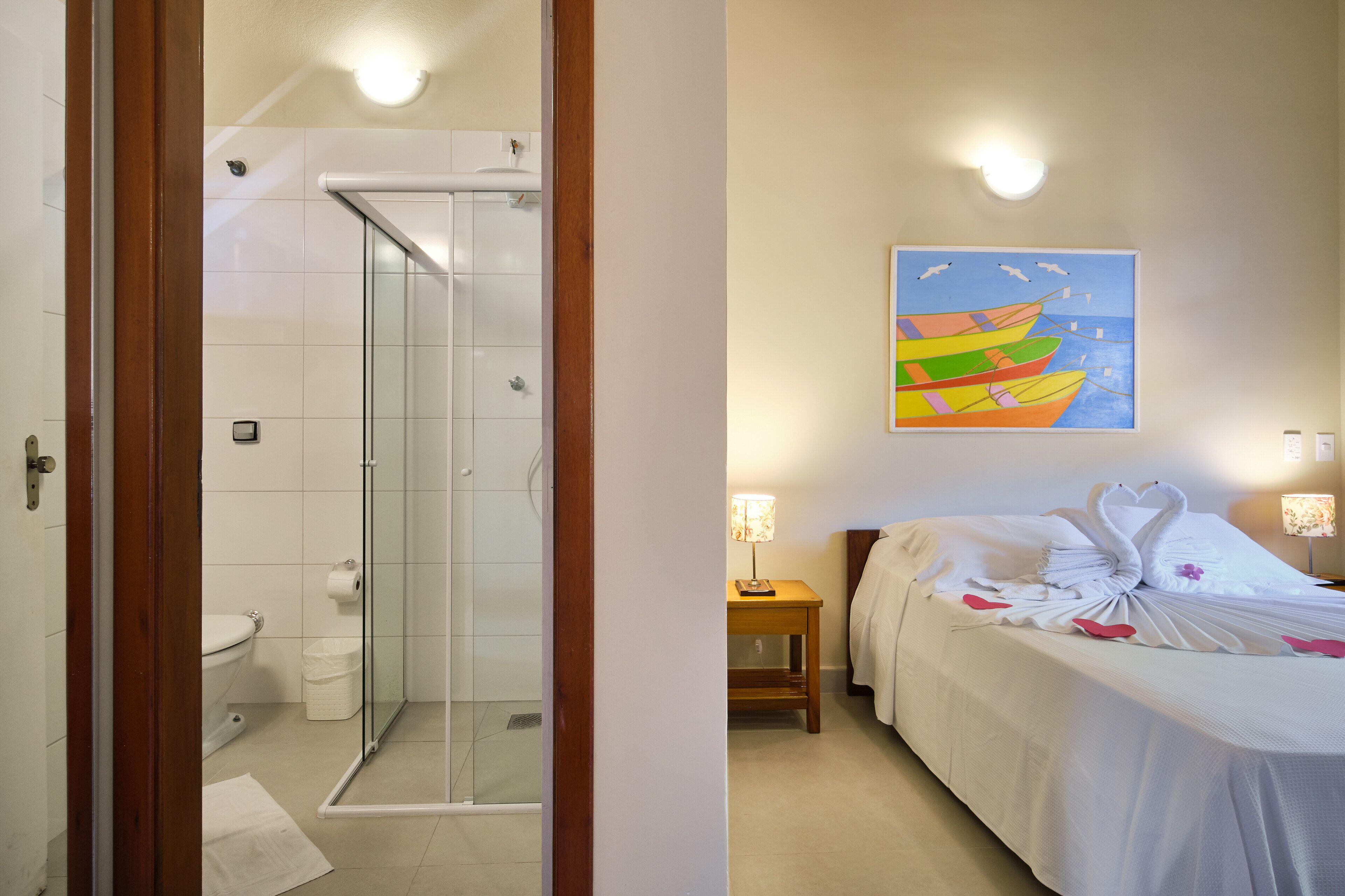 A bathroom with a glass shower enclosure and a toilet. There is a bed with white bedding and a decorative pillow arrangement.