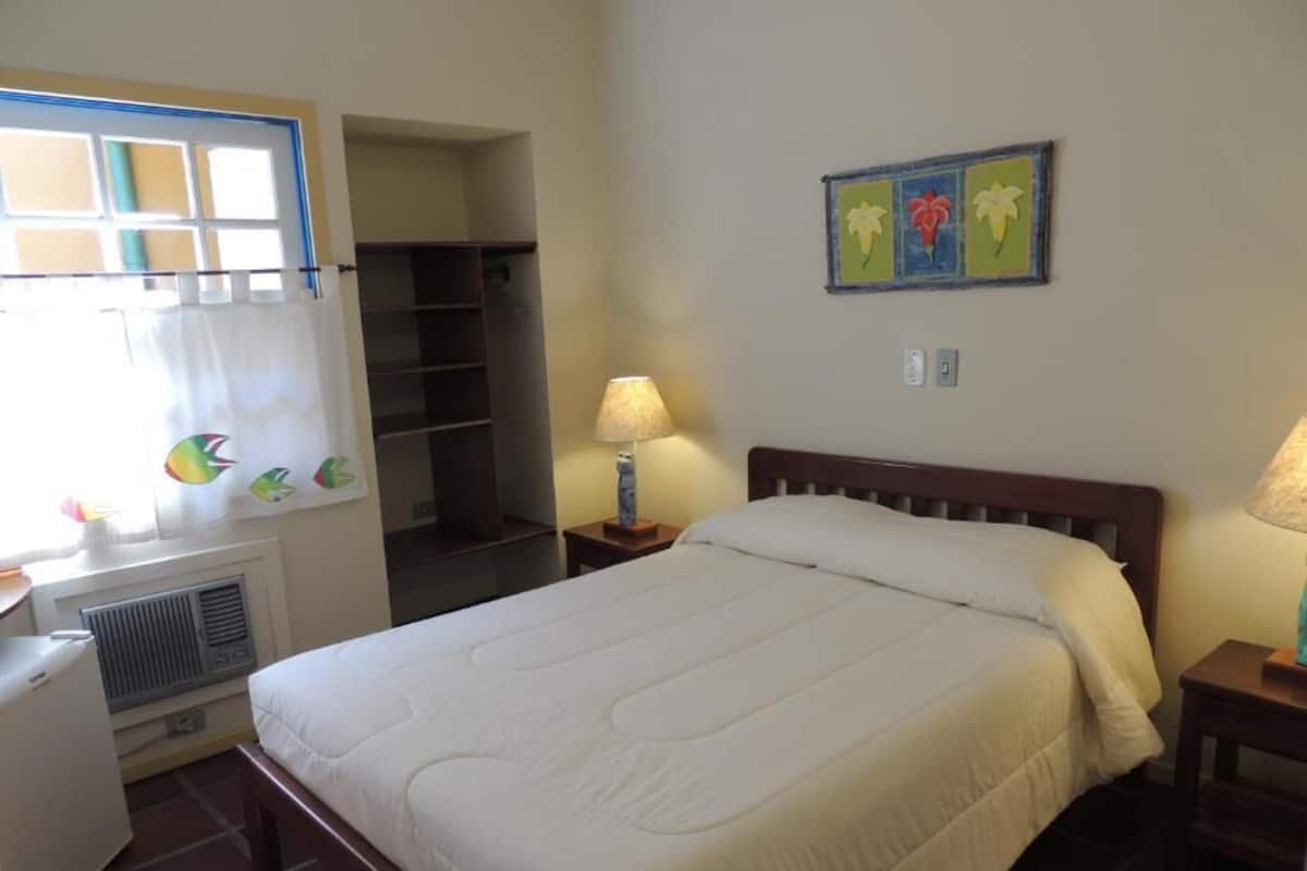 Un dormitorio con cama, mesitas de noche, lámpara, calefactor, armario y ventana con cortinas.