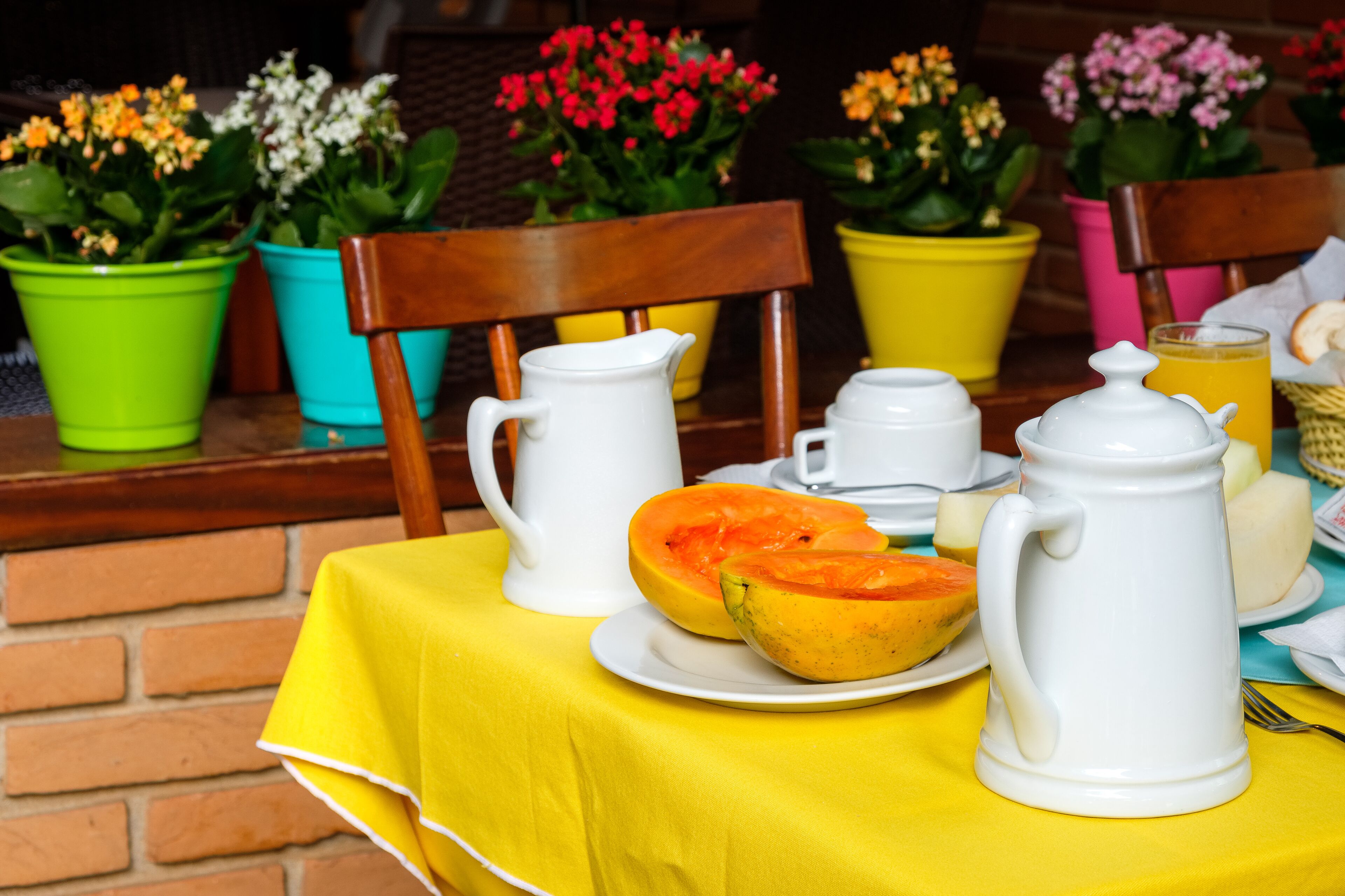 Une table de petit-déjeuner dressée avec une nappe jaune, une cafetière et une théière en céramique blanche, et une demi-papaye coupée sur une assiette.