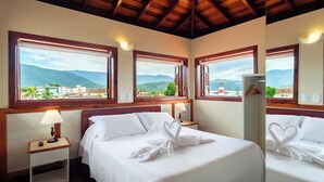 A hotel room with a bed, two bedside tables with lamps, and large windows offering a view of mountains and buildings. - Pousada Don Diego (Ubatuba)