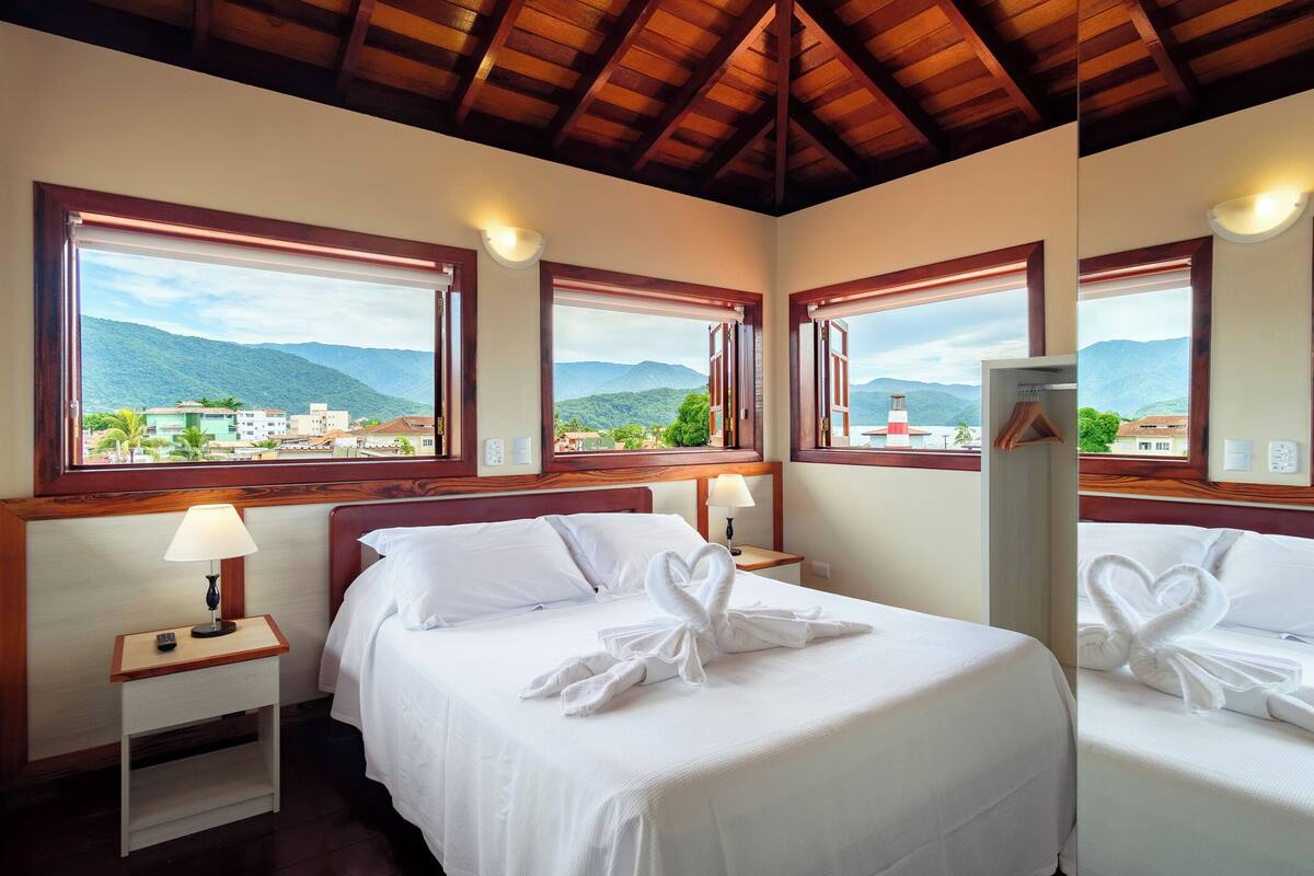 Habitación de hotel con cama, dos mesitas de noche con lámparas y ventanas amplias que ofrecen vistas a montañas y edificios.