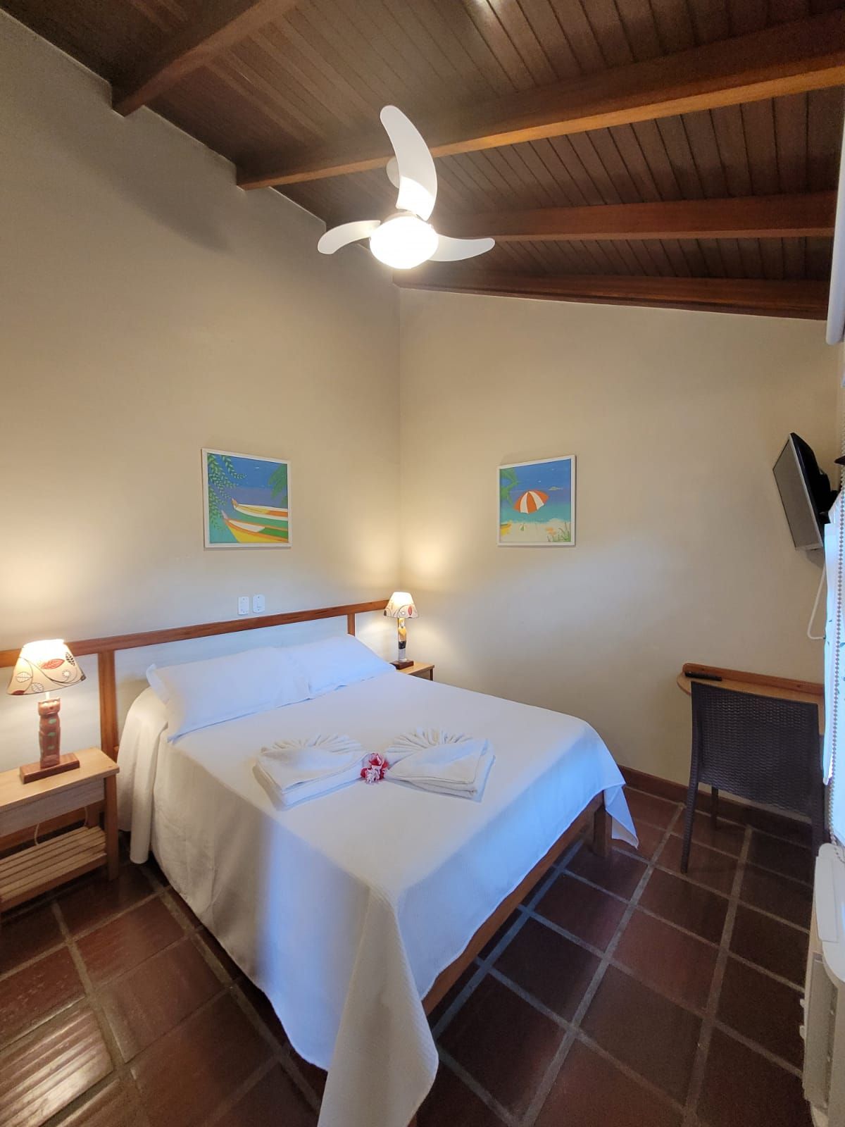 Una habitación de hotel con una cama, dos lámparas de noche, un ventilador de techo y dos cuadros colgados en la pared.