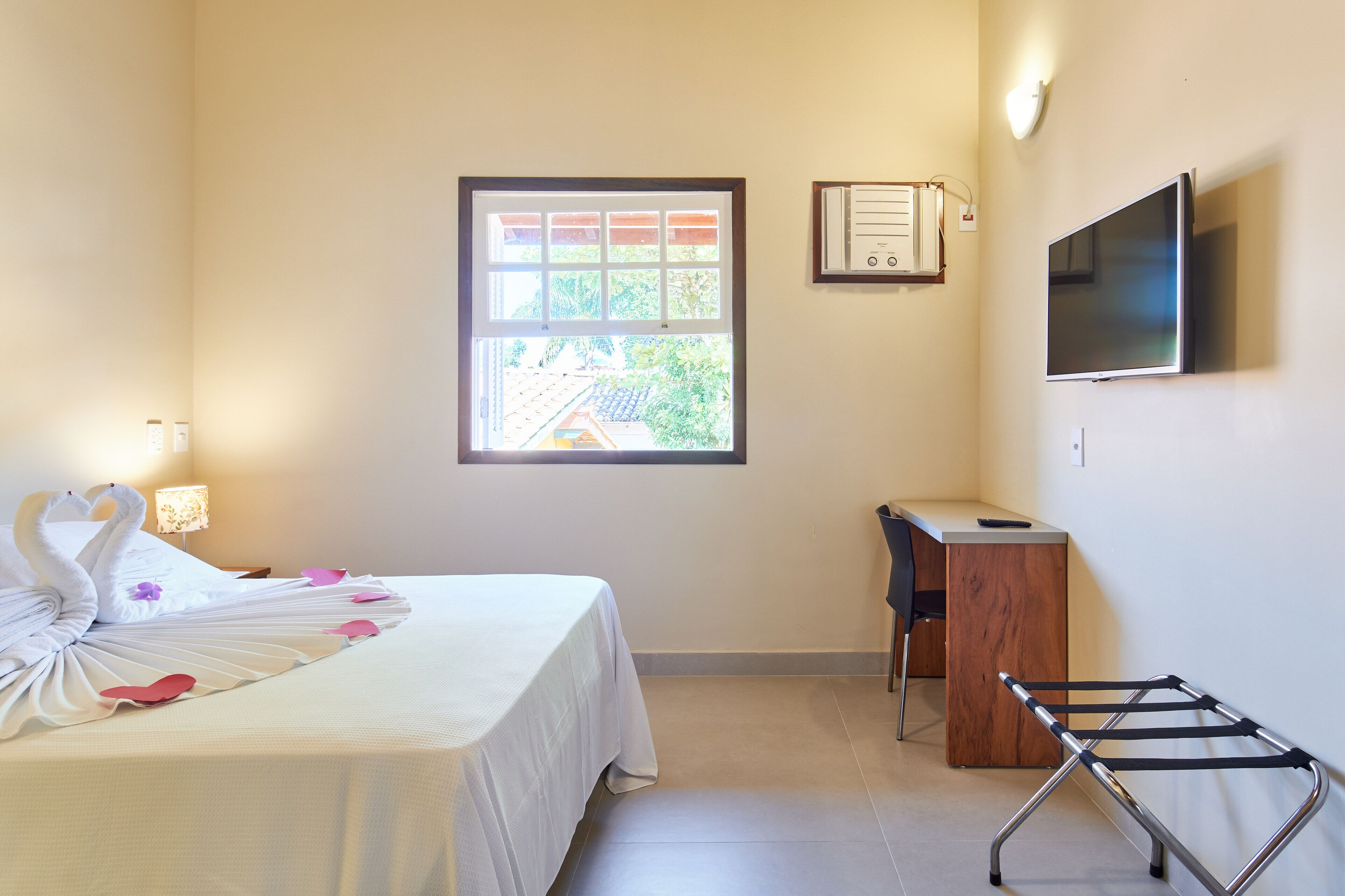 A hotel room with a bed, a desk, a TV, a window, and a wall-mounted air conditioning unit.