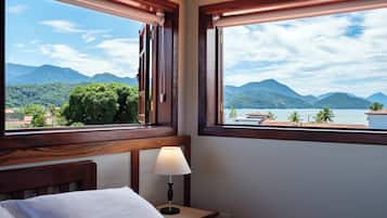 A hotel room with a bed, a nightstand with a lamp, and a window offering a view of mountains and a lake.