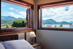 A hotel room with a bed, a nightstand with a lamp, and a window offering a view of mountains and a lake. - Pousada Don Diego (Ubatuba)