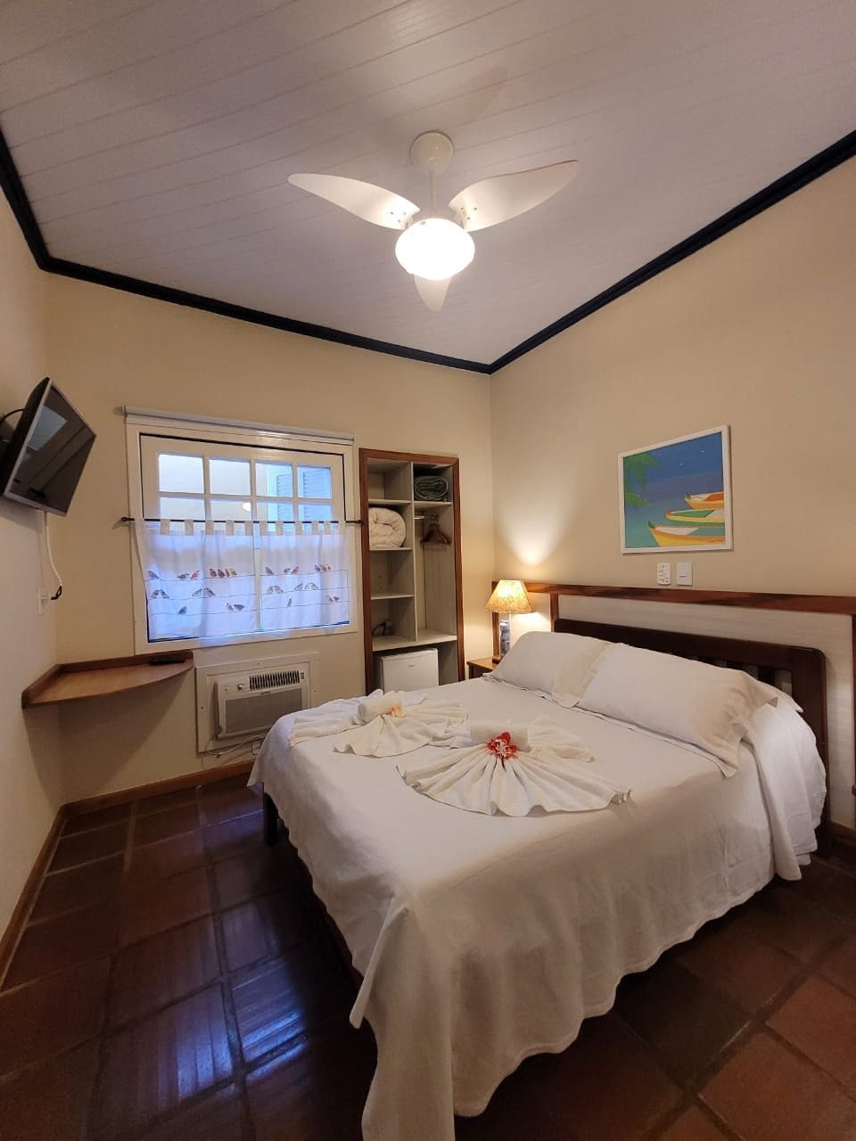 Une chambre d’hôtel avec un lit, un ventilateur de plafond, une fenêtre avec des rideaux, un tableau au mur et un téléviseur fixé au mur.