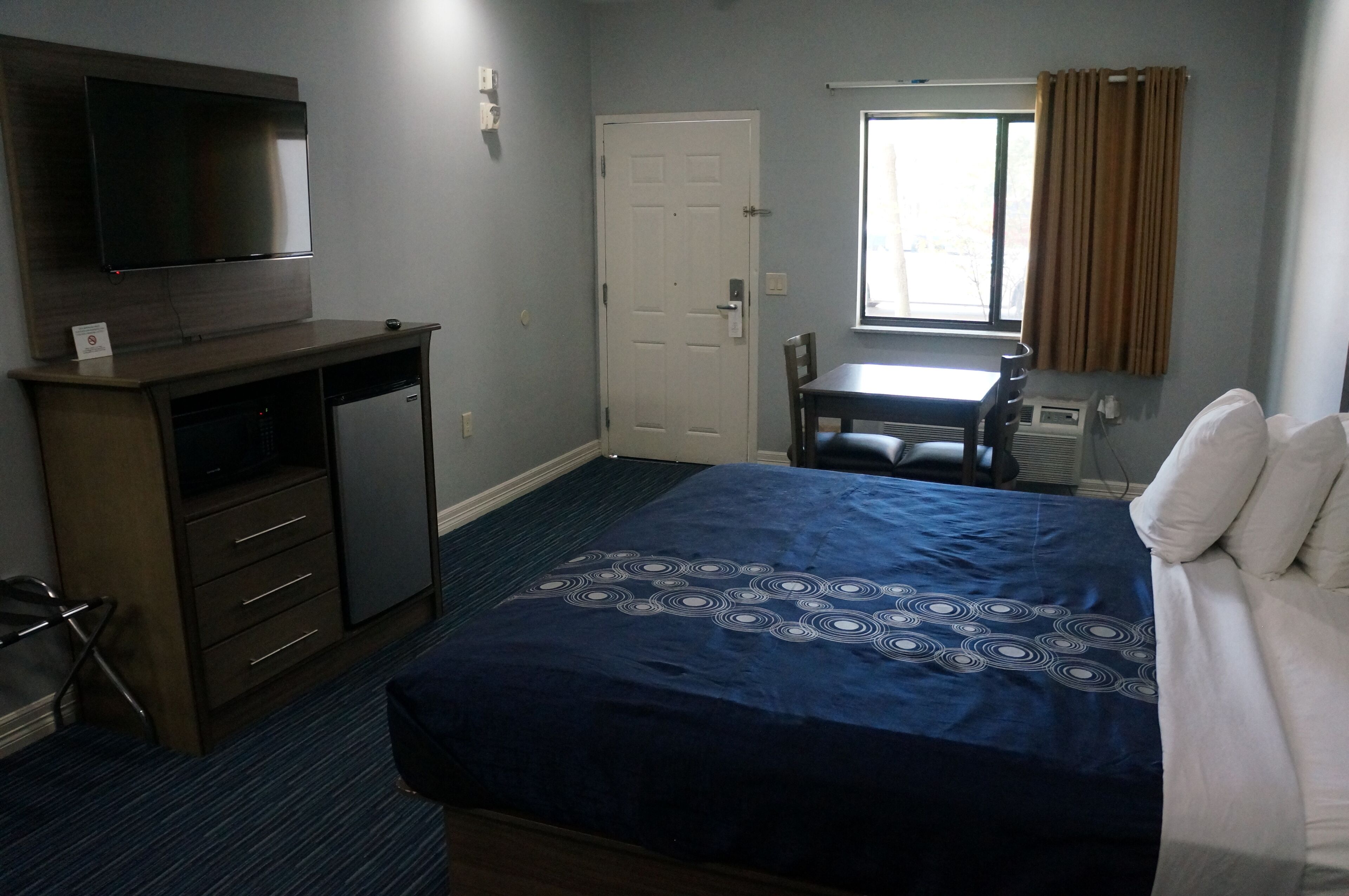 A hotel room with a bed, a desk with a computer, a television, a window with curtains, and a door.