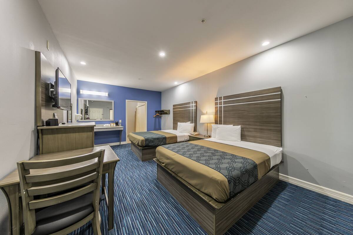 A hotel room with two beds, a desk, a chair, a mirror, and a bathroom with a sink and mirror.