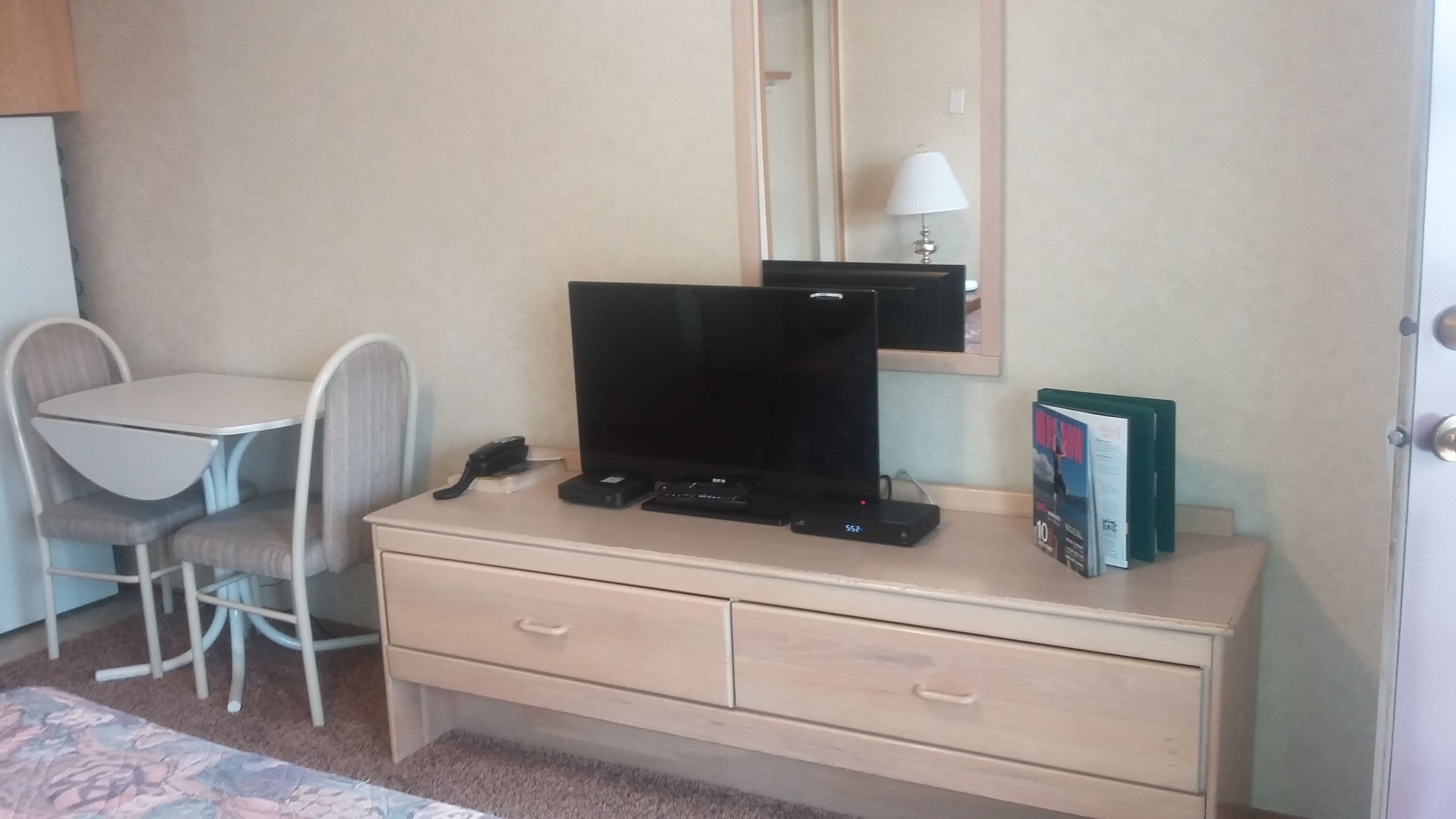 Habitación de hotel con escritorio, una silla, televisor, lámpara y una revista sobre el escritorio.