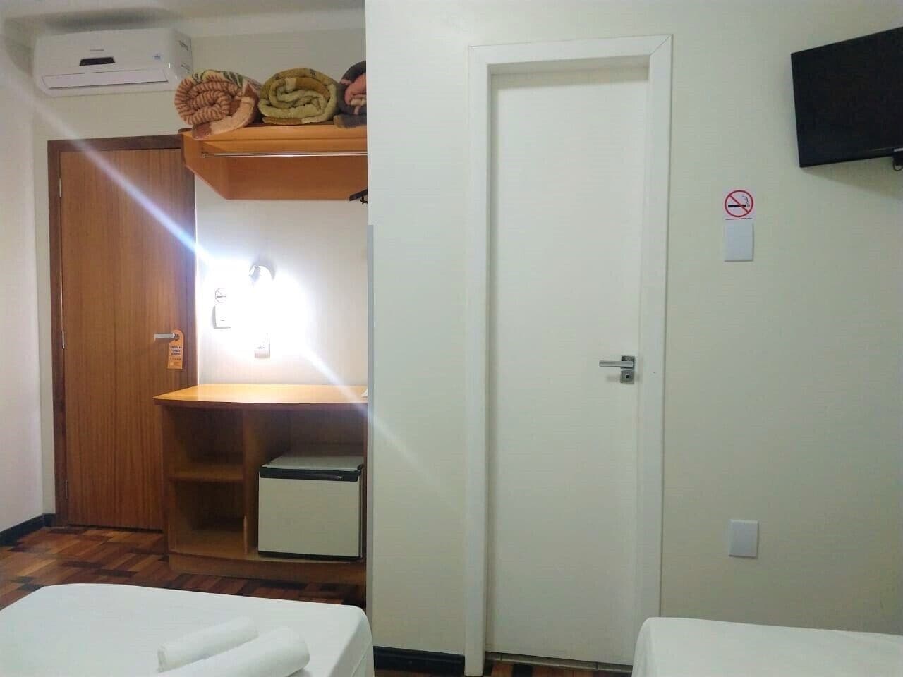 Ein Hotelzimmer mit einem Holfschrank, einem kleinen Schreibtisch mit Lampe, einer weißen Tür und einem an der Wand befestigten Fernseher.