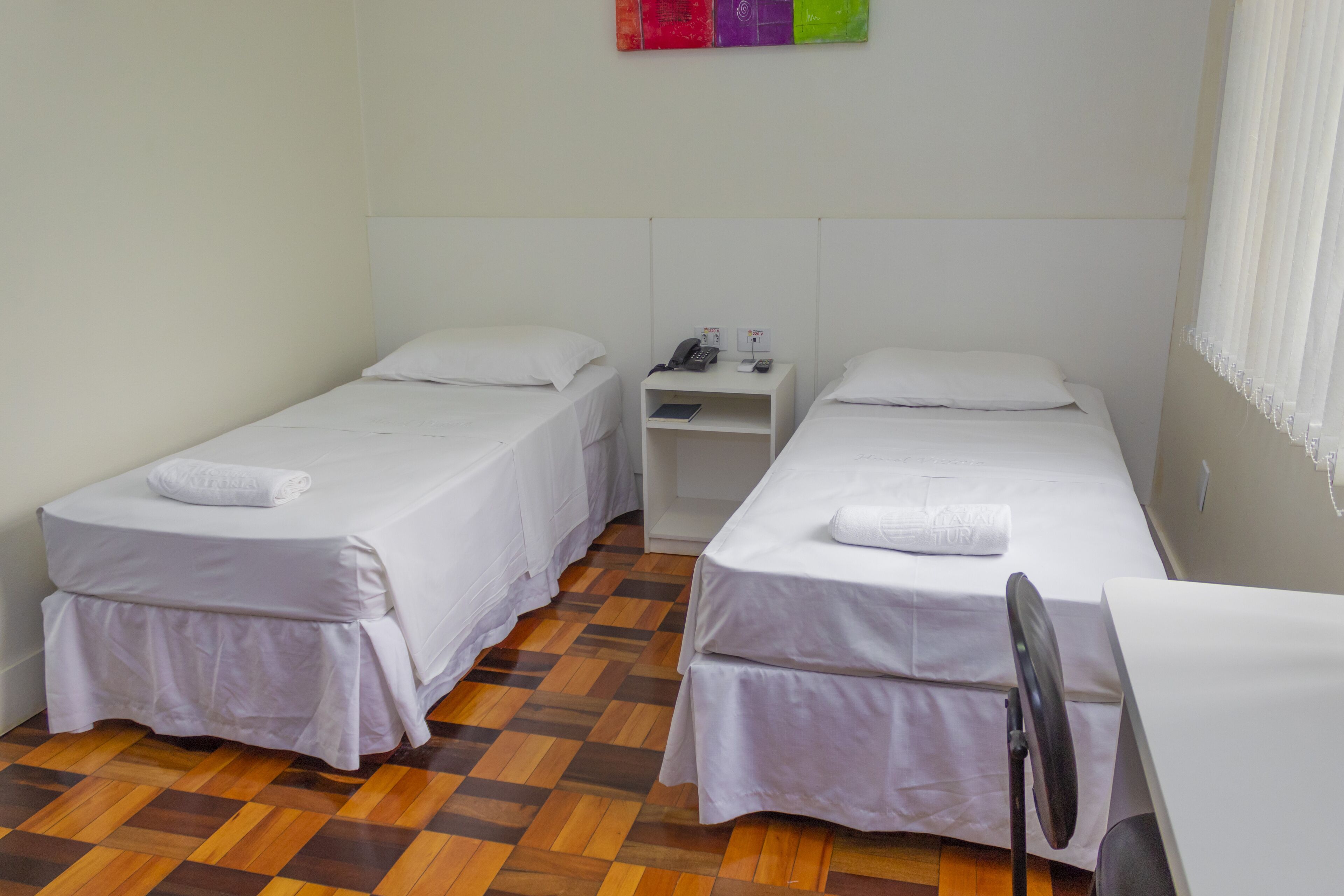 Habitación de hotel con dos camas individuales, un escritorio pequeño, una silla y una mesita de noche con teléfono y un libro.