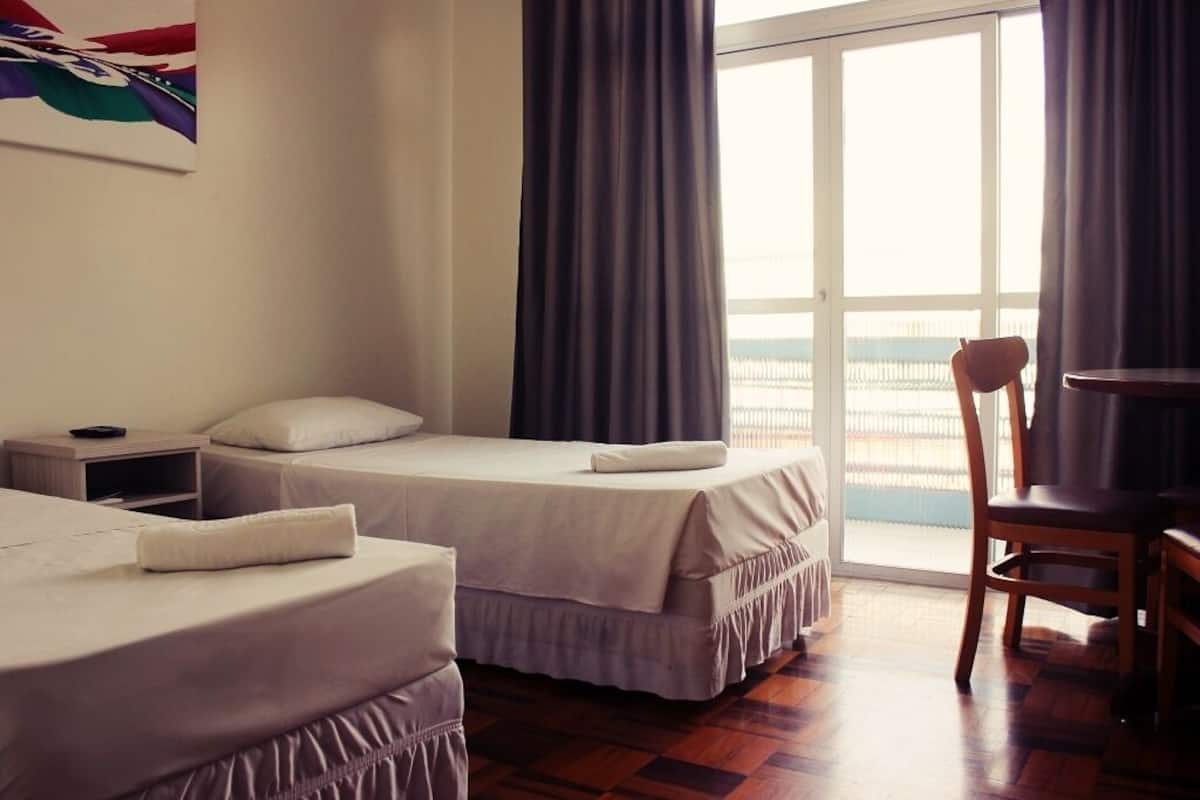Habitación de hotel con dos camas, una silla de madera, una mesita y vistas al mar a través de un ventanal grande.
