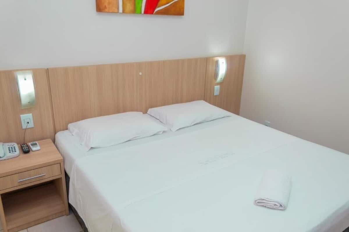Habitación de hotel con cabecera de madera, cama individual con ropa blanca, mesita de noche con teléfono y control remoto, y lámpara fijada a la pared.