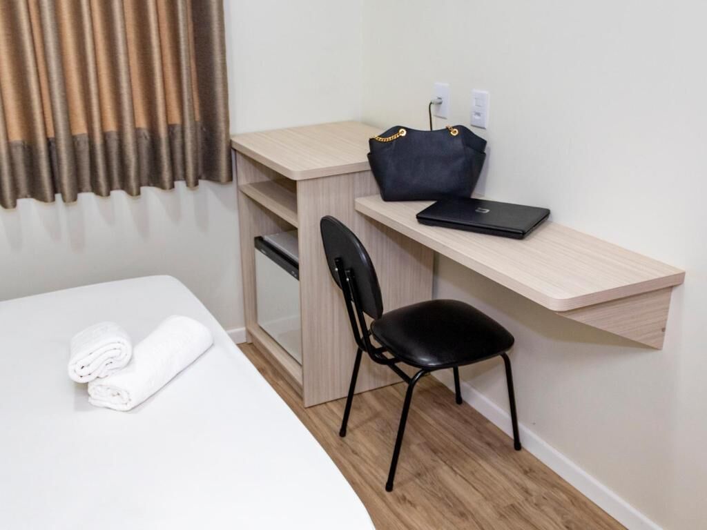A hotel room with a bed, a desk with a chair, a laptop, a handbag, and a rolled-up towel.