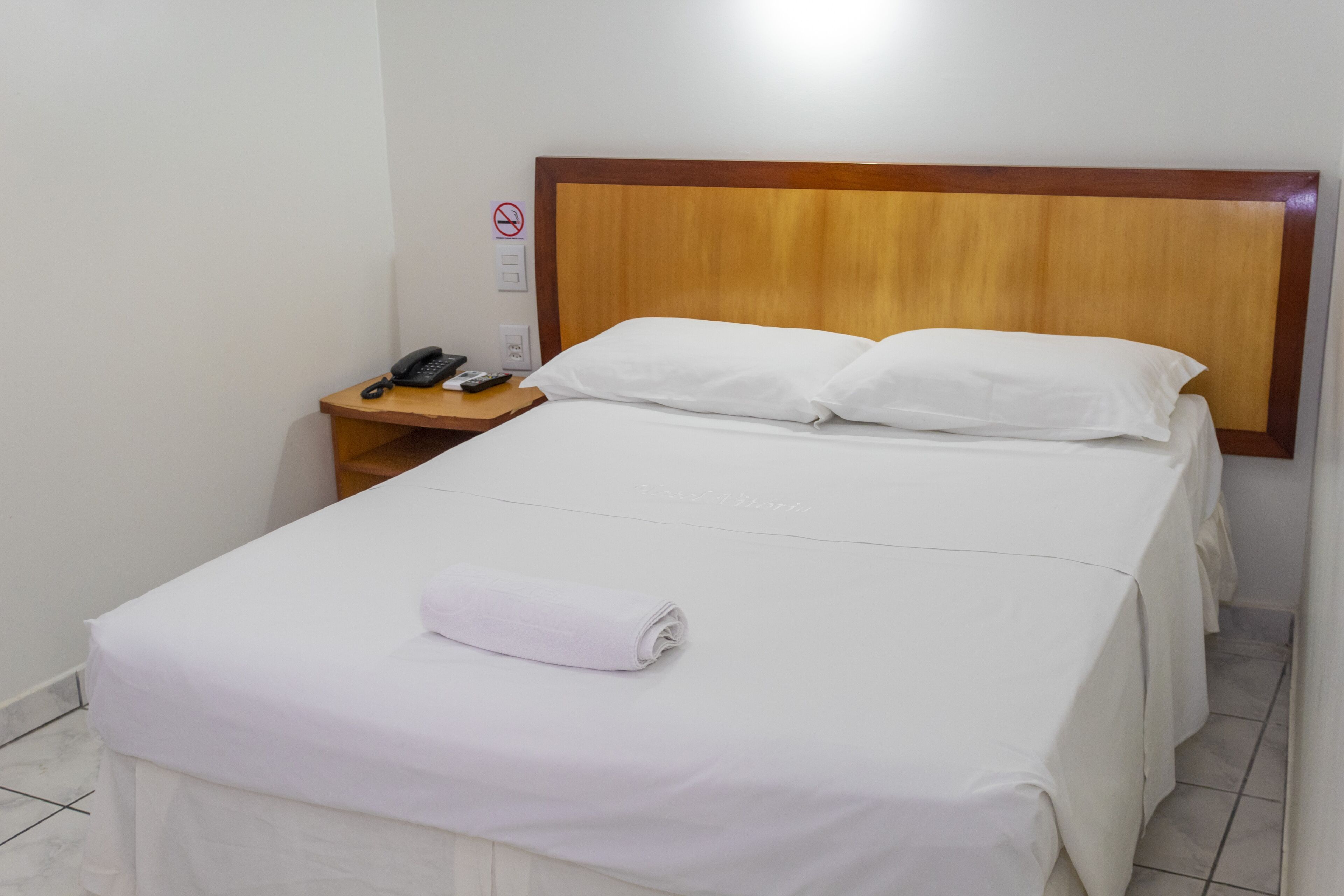 A hotel room with a neatly made bed, a bedside table with a phone and remote control, and a no-smoking sign on the wall.