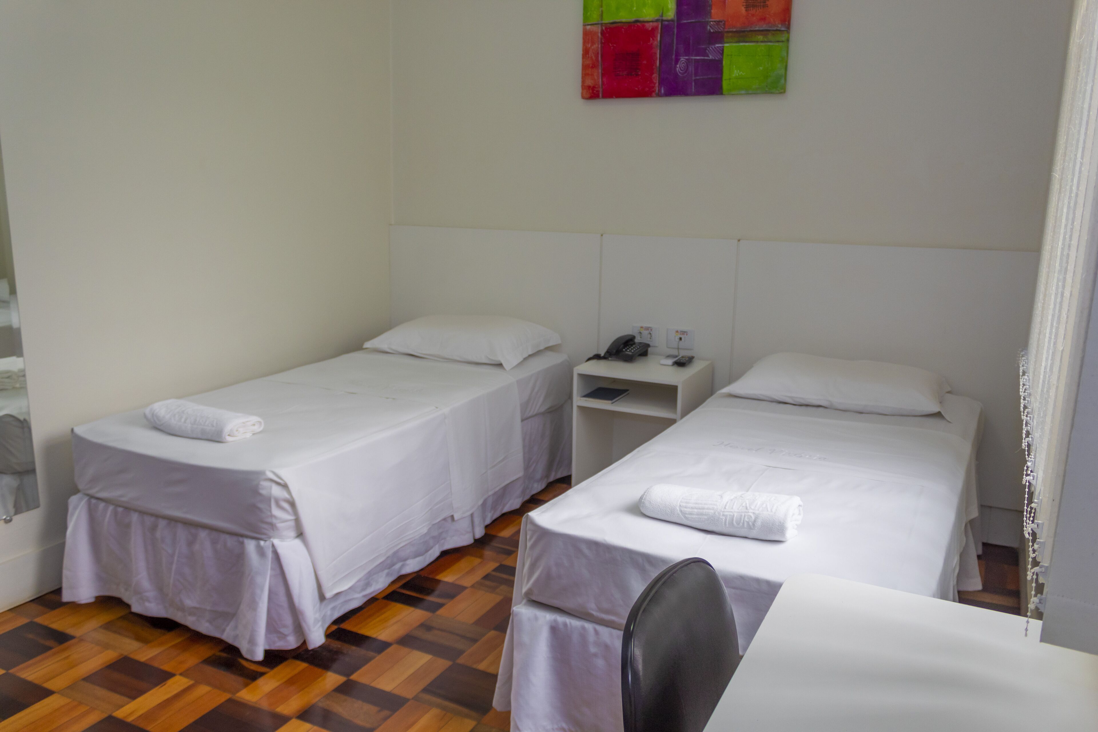 Habitación de hotel con dos camas individuales, una mesita de noche pequeña con un teléfono y un cuadro abstracto colorido en la pared.