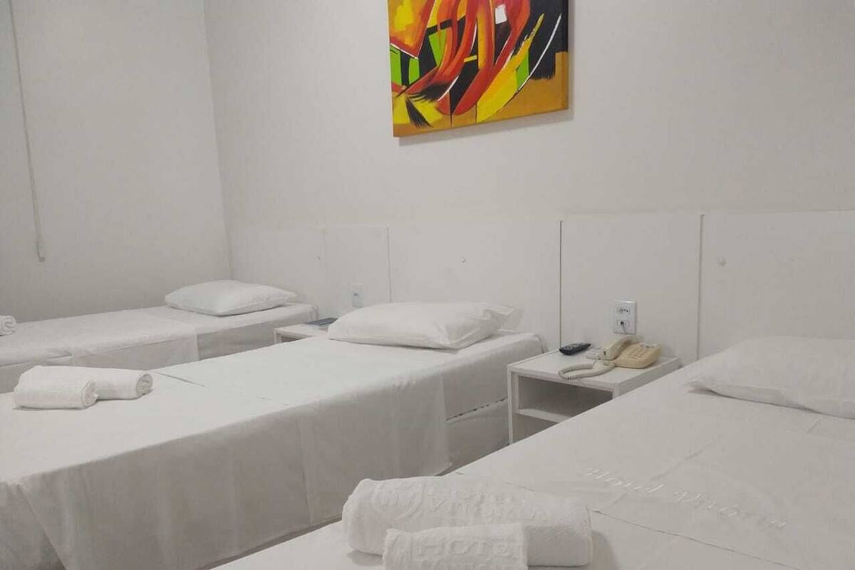 Habitación de hotel con tres camas individuales, cada una con cubrecama y almohada blancos, una mesita de noche con teléfono, y un cuadro abstracto de colores en la pared.