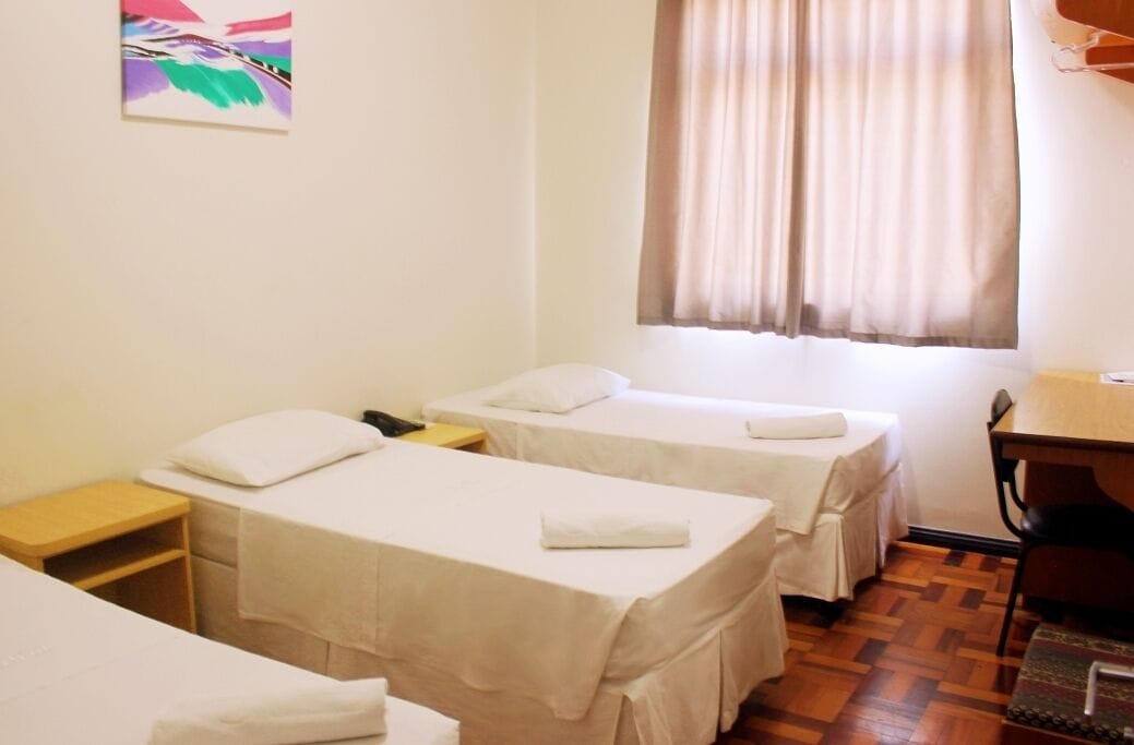 A hotel room with two single beds, a wooden desk, a chair, and a window with curtains.