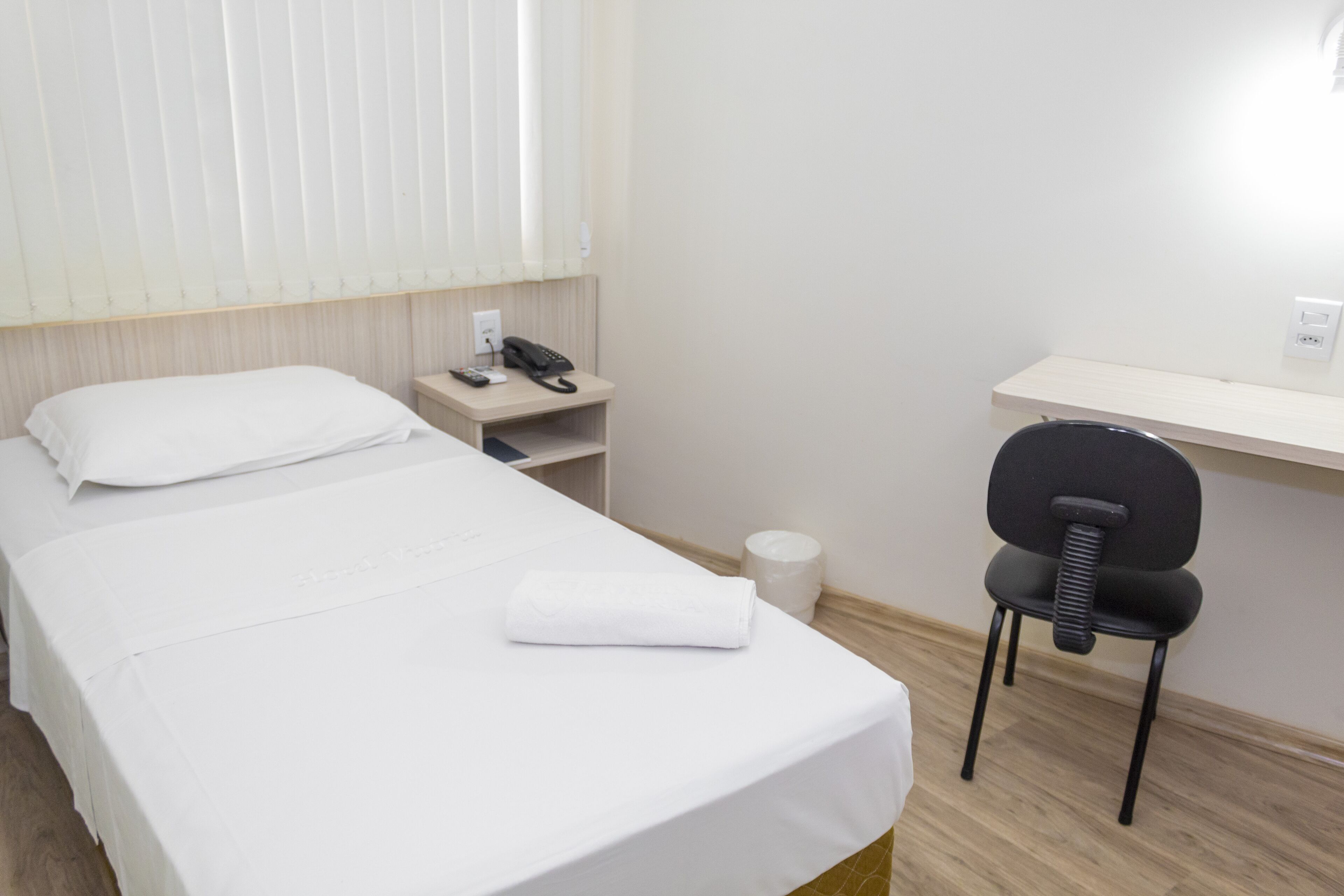 A hotel room with a neatly made bed, a desk with a chair, a telephone, and a waste bin.
