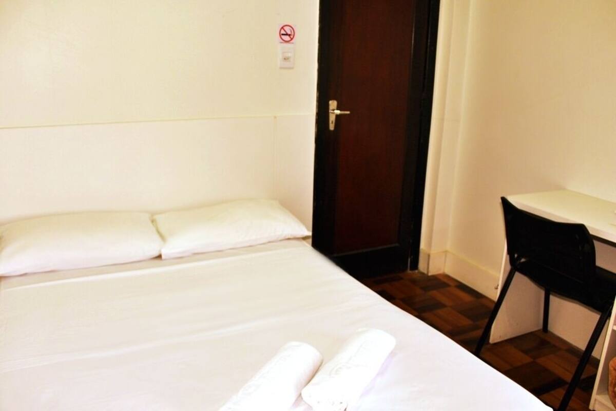 Una cama individual con ropa blanca, una puerta de madera, un escritorio pequeño con una silla y una pared blanca con un cartel de prohibido fumar.