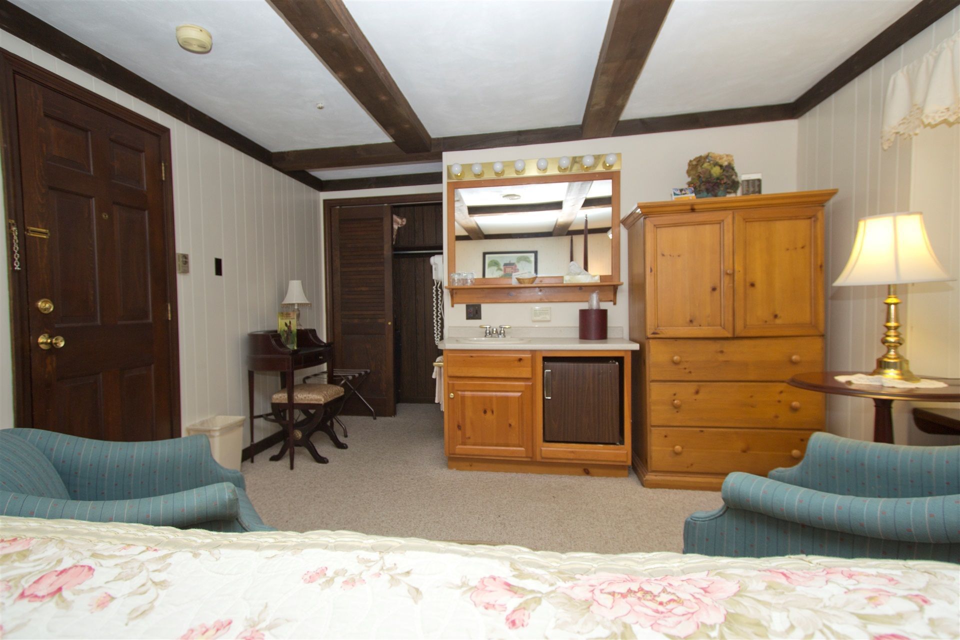 A hotel room with a bed, a desk with a lamp, a wardrobe, and a mirror with lights.