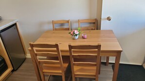 Una mesa de comedor de madera con cuatro sillas, un pequeño jarrón con flores y un refrigerador al fondo.