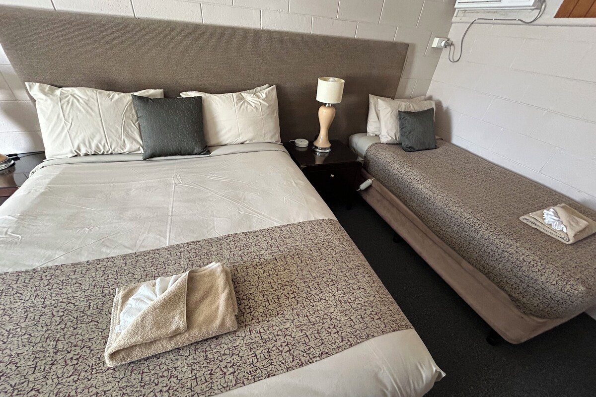 A hotel room with a large bed, a smaller bed, a nightstand with a lamp, and a folded towel on the floor.
