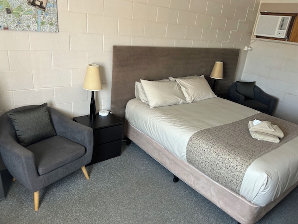 Habitación de hotel con cama, dos mesitas de noche, una silla, una lámpara y aire acondicionado instalado en la pared.