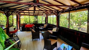 An open-plan lounge with a high ceiling and wooden beams, furnished with wicker chairs and tables, and large windows offering views of greenery outside. - Hotel Belvedere (Sámara)