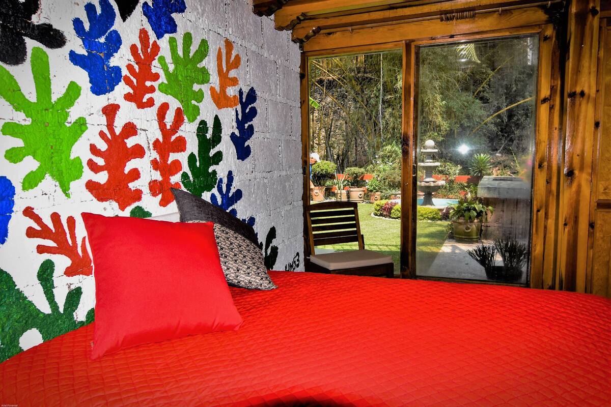 Een slaapkamer met een rode sprei, een kleurrijke muurschildering, een houten deur die naar een tuin leidt, en een klein zithoekje met een stoel en een bankje.