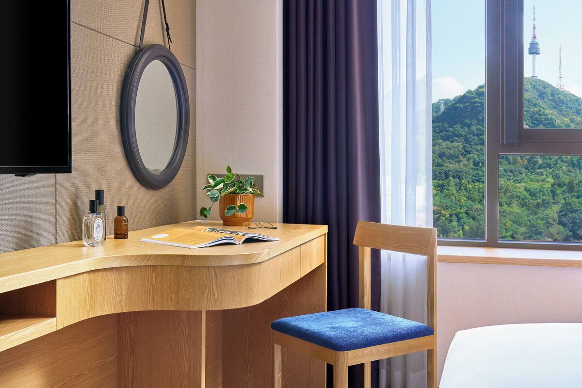 A hotel room with a wooden desk, a chair, a bed, a TV, a mirror, a window with a view of a mountain, and a potted plant.