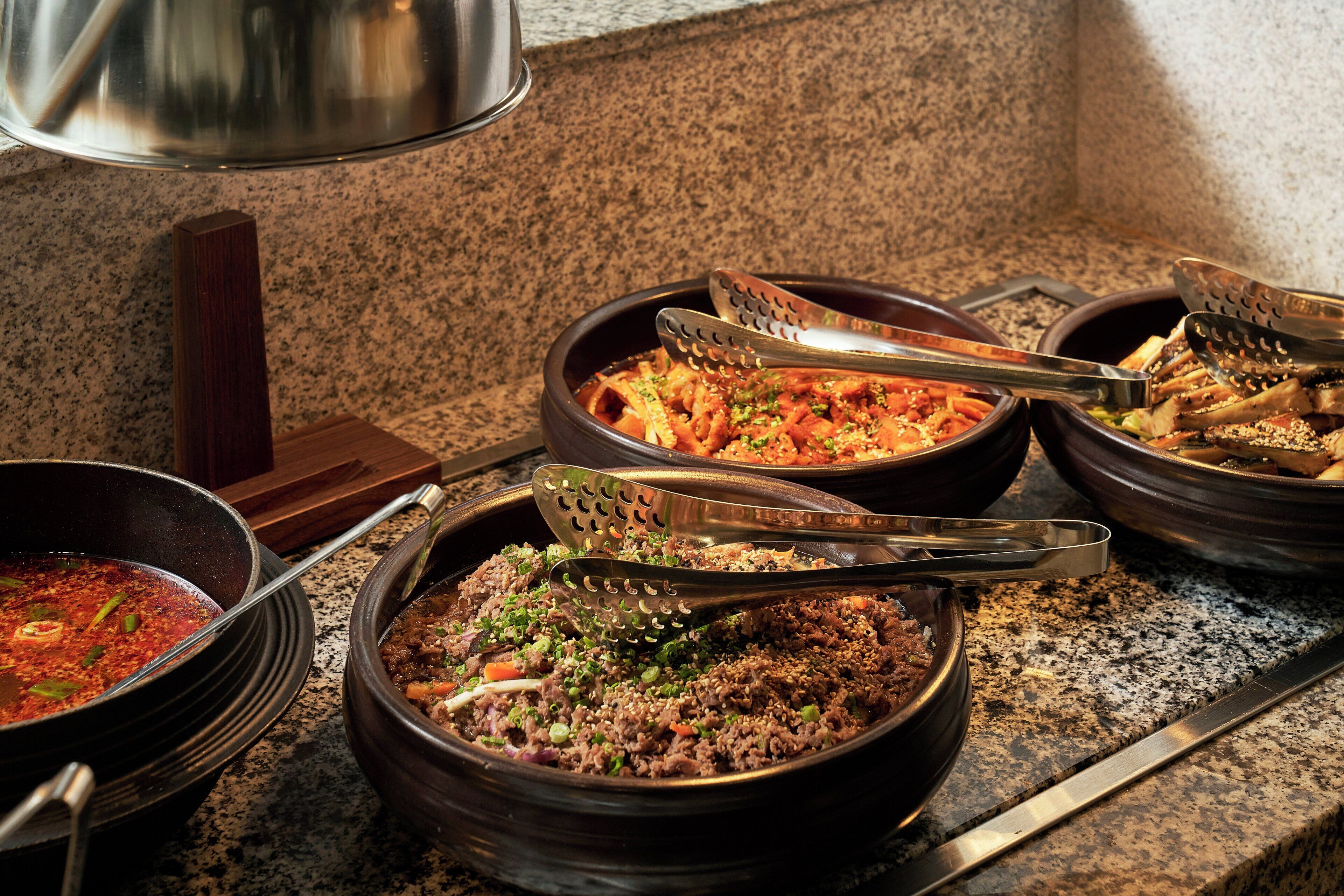 A variety of dishes served in traditional black bowls with metal tongs on a granite countertop.