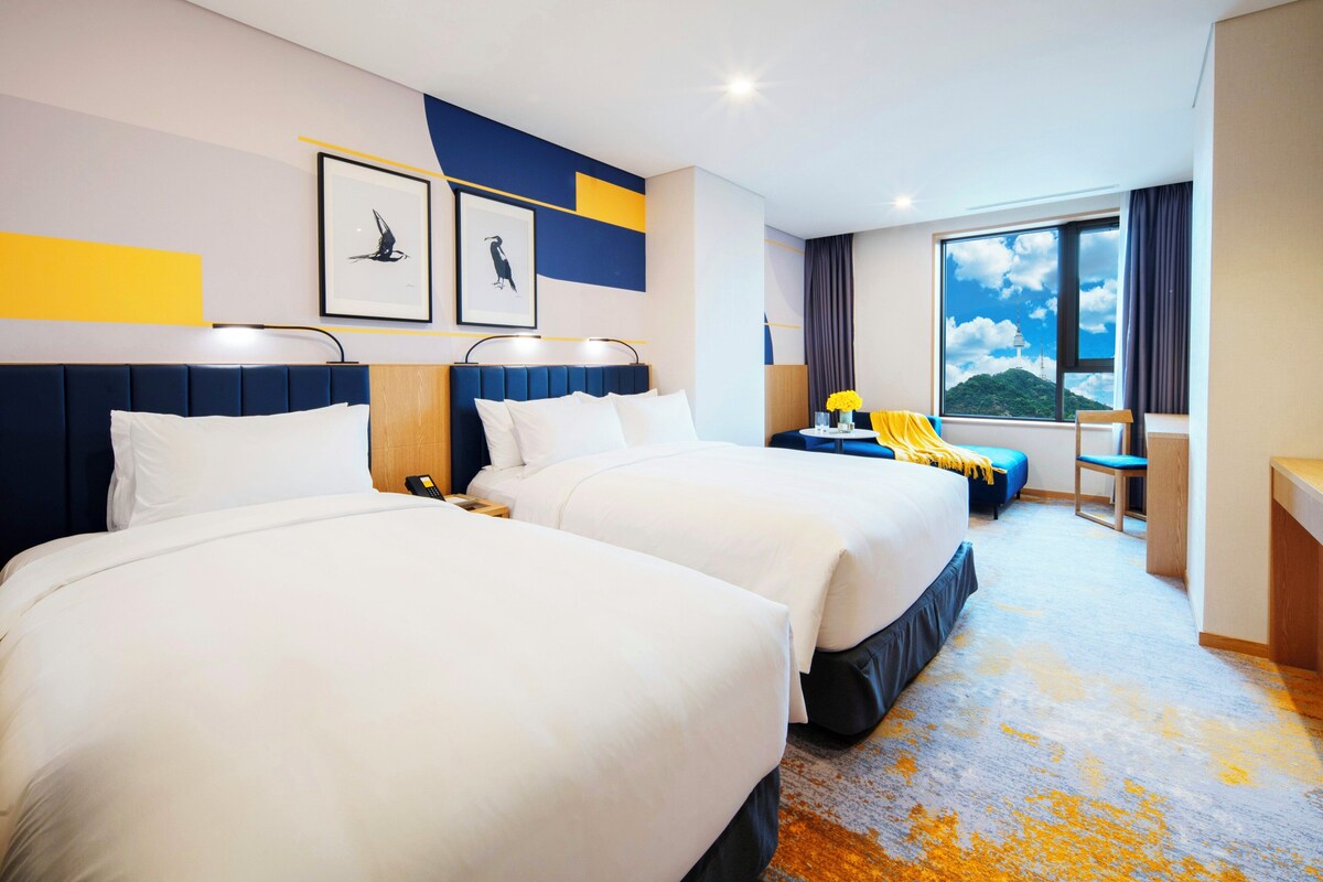 A hotel room with two beds, a desk, a chair, a window with a view, and artwork on the wall.