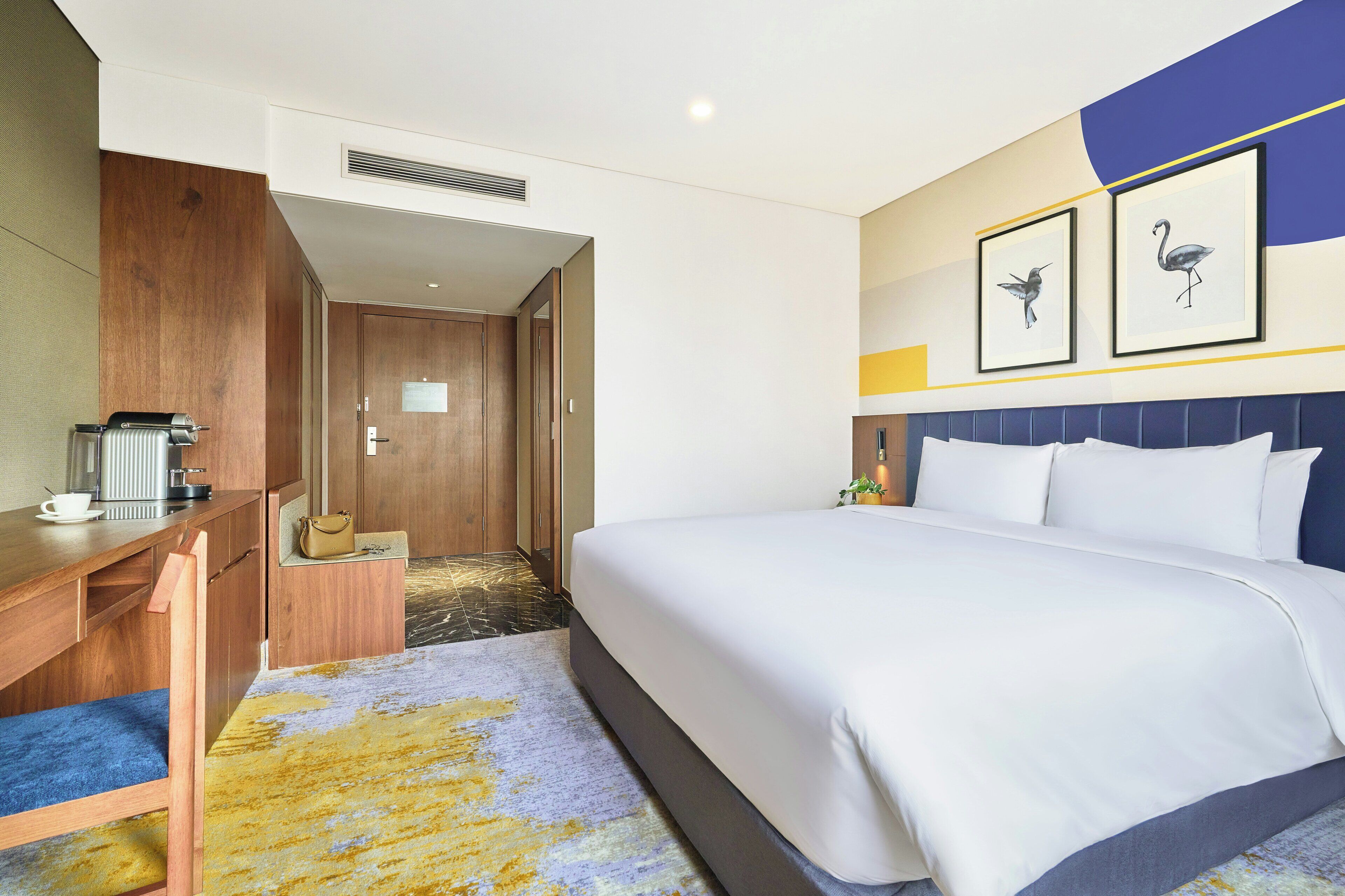 Une chambre d’hôtel avec un lit, un bureau, une chaise, une machine à café et un tableau encadré au mur.