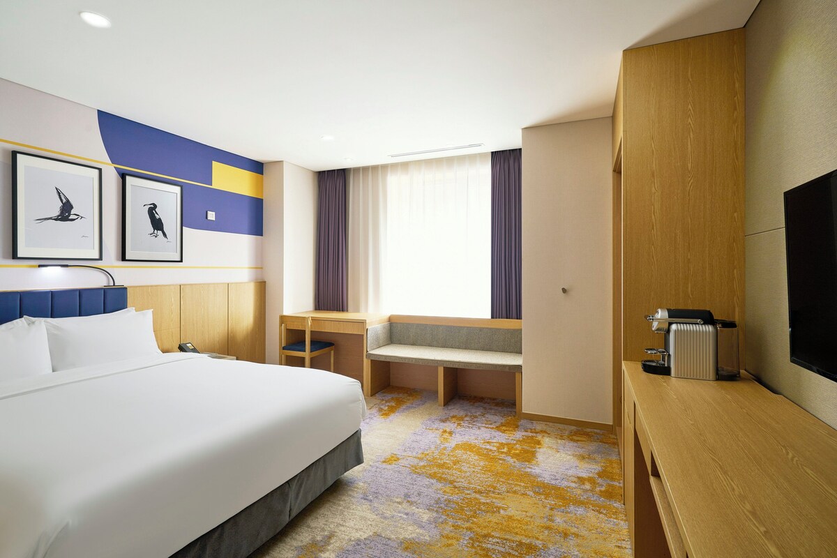 A hotel room with a bed, a desk, a chair, a television, and artwork on the wall.