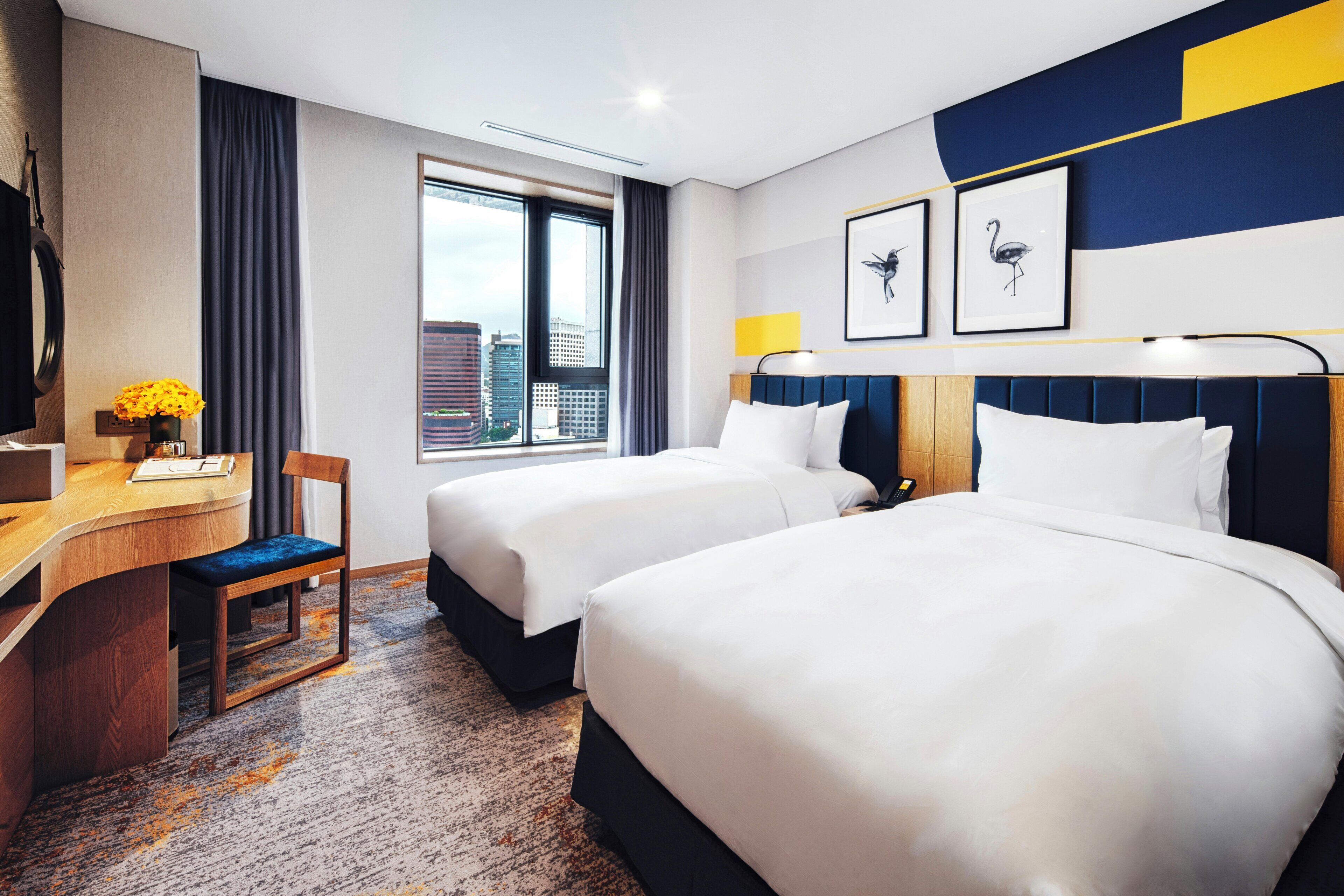 A hotel room with two beds, a desk with a chair, a window with a city view, and artwork on the wall.