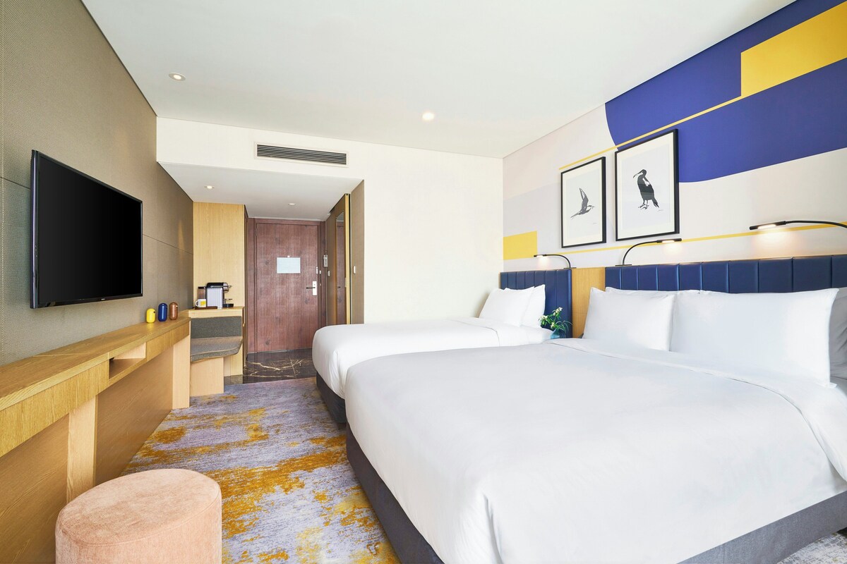 A hotel room with a bed, a desk with a flat-screen TV, a coffee machine, and a door leading to another room.